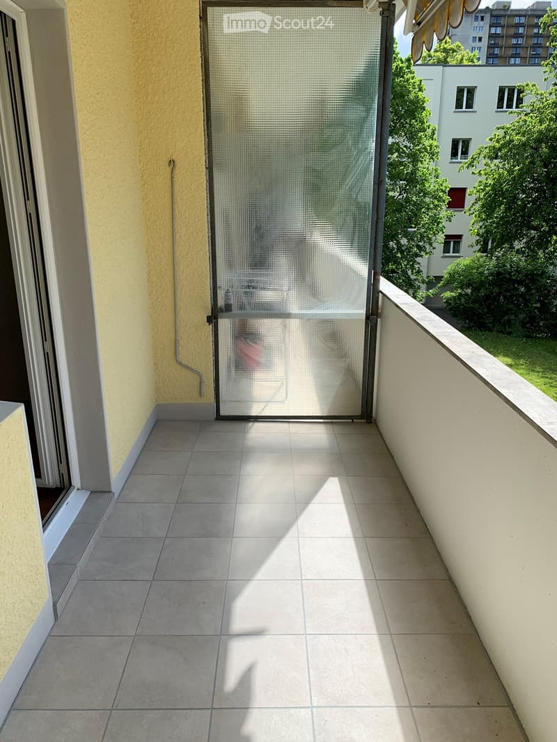 Balcony with tiled floor, glass sliding door, and metal railing, overlooking a green area with trees and buildings.