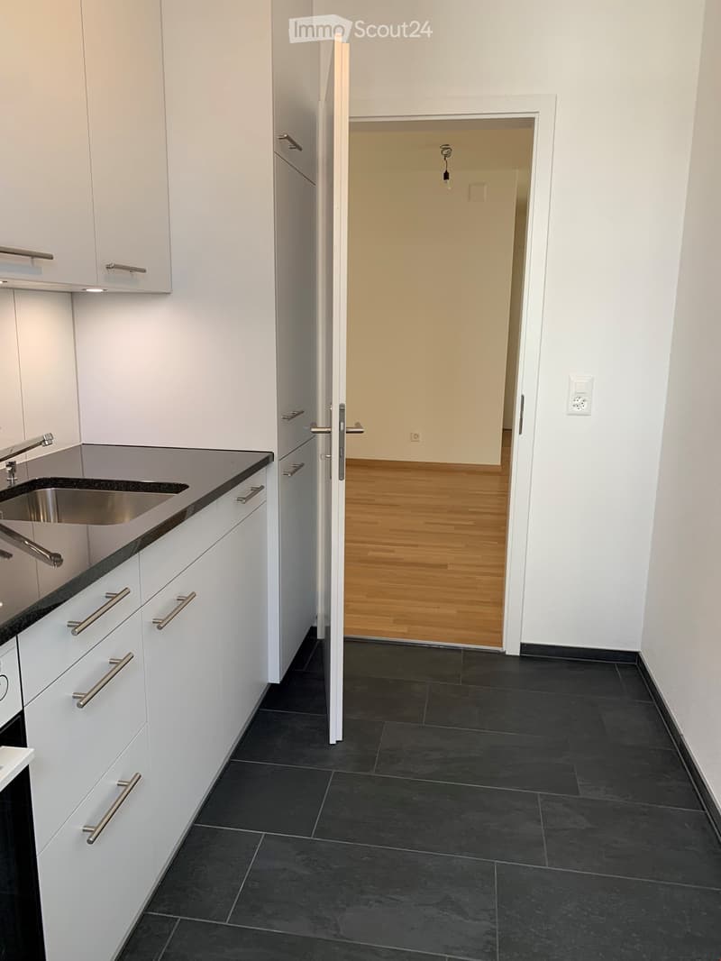 Modern kitchen with white cabinets, black countertops, and a stainless steel sink. The floor is dark tiled, and an open doorway leads to another room with wooden flooring.