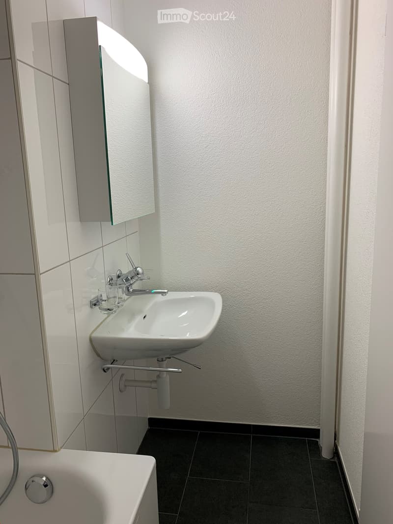 Modern bathroom with white tiled walls, a white sink with a chrome faucet, a mirror cabinet, a glass cup on the sink, and a bathtub with a chrome handle.
