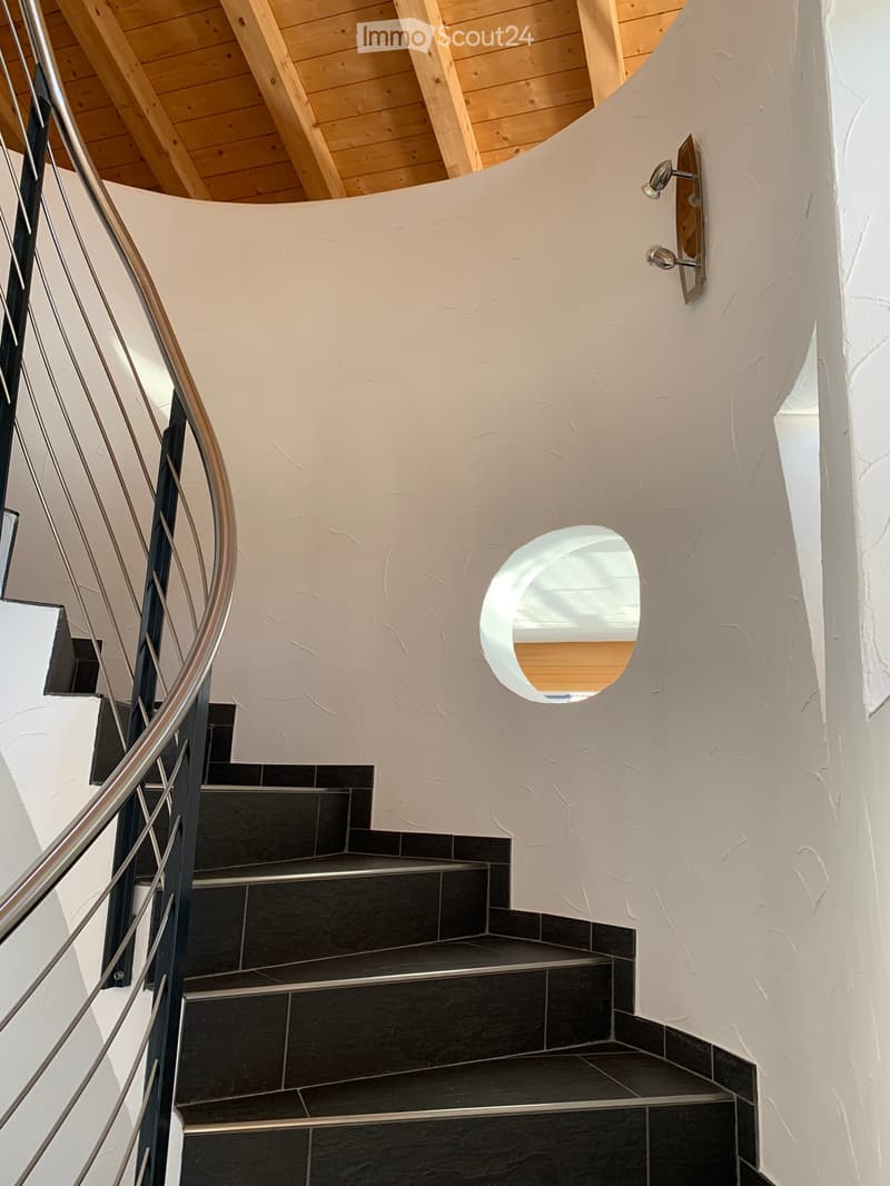 Spiral staircase with dark tiled steps and a modern metal railing, featuring a textured white wall and a circular window at the top.