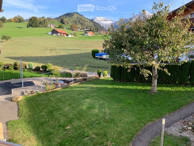 Well-maintained garden with green lawn, gravel pathways, small water feature, and surrounding trees. Scenic mountain view in the background.