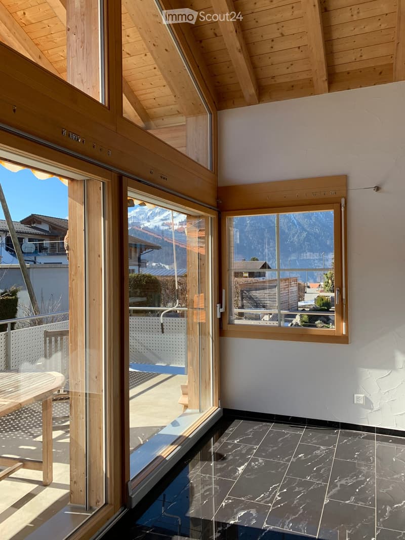 Bright room with wooden beams, large windows offering mountain views, black tiled floor, and a wooden table near the window.