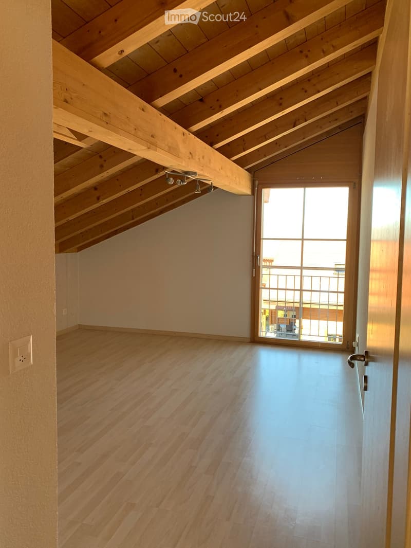 Empty attic space with wooden beams, wooden flooring, and a large window leading to a balcony.