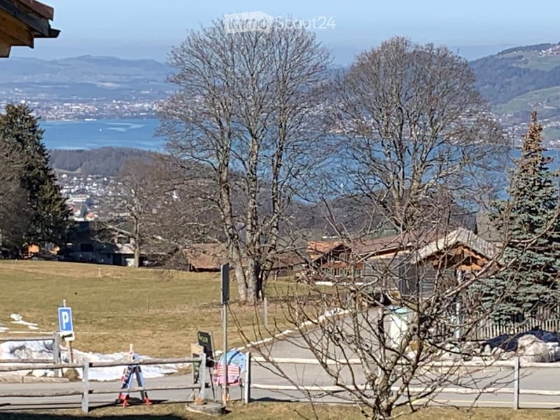 View of a scenic outdoor area with a lake, mountains, and scattered houses in the background. The foreground includes a wooden fence, a parking sign, and some bare trees.