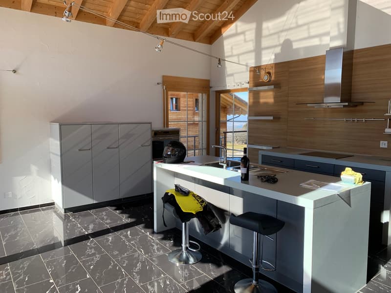 Modern kitchen with grey cabinets, island, sink, and stools. Features a sleek design with wooden accents and a view through large windows.
