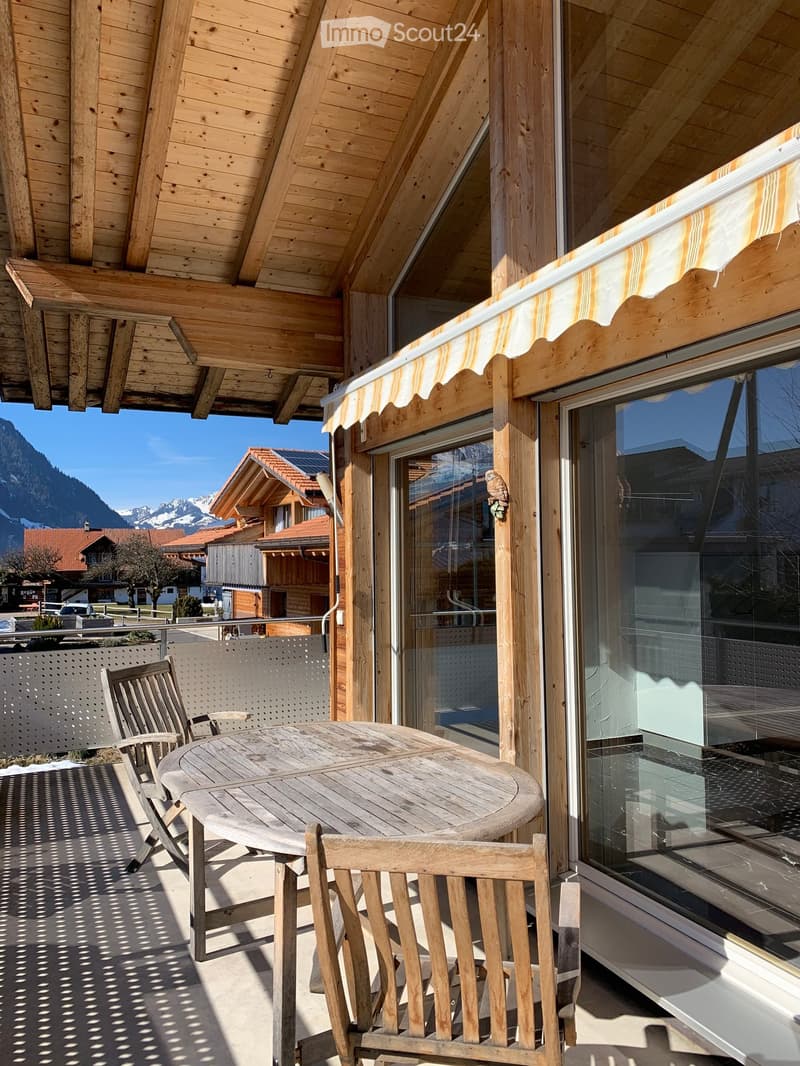Wooden balcony with a round table and chairs, overlooking a scenic mountain view.
