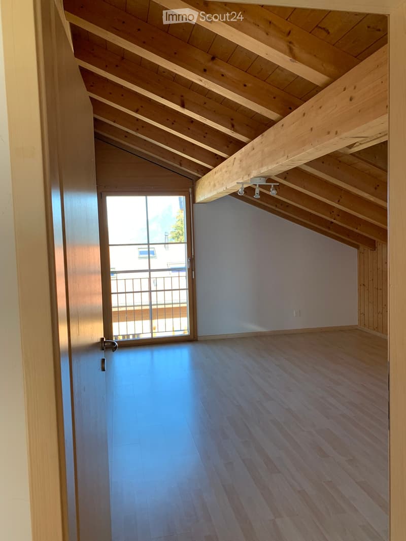 Empty attic space with wooden beams, wooden floor, and a large window with a metal railing visible outside.