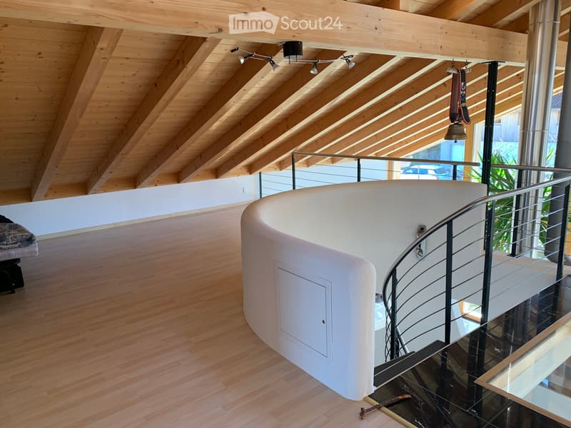 Spacious hallway with wooden flooring, exposed wooden ceiling beams, modern lighting fixtures, and a curved white wall with a built-in storage compartment. A modern metal railing leads to an upper level with glass balustrades and visible greenery outside.
