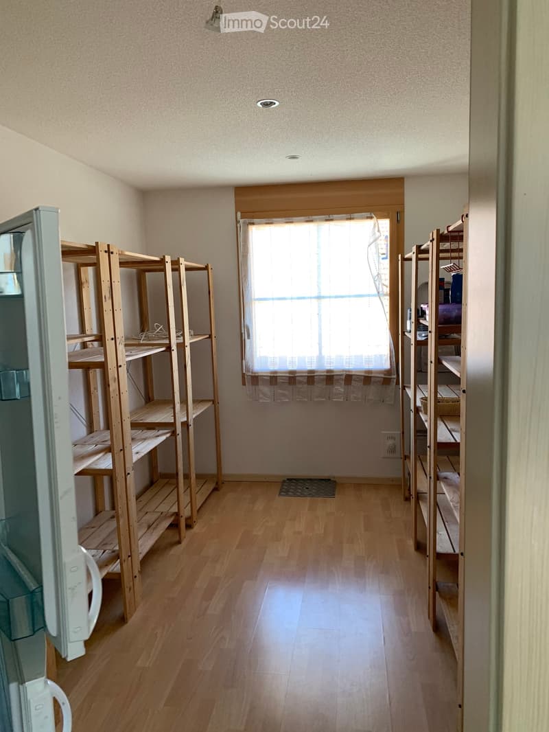 A room with wooden shelves on both sides, a window with a curtain, and a refrigerator on the left side.