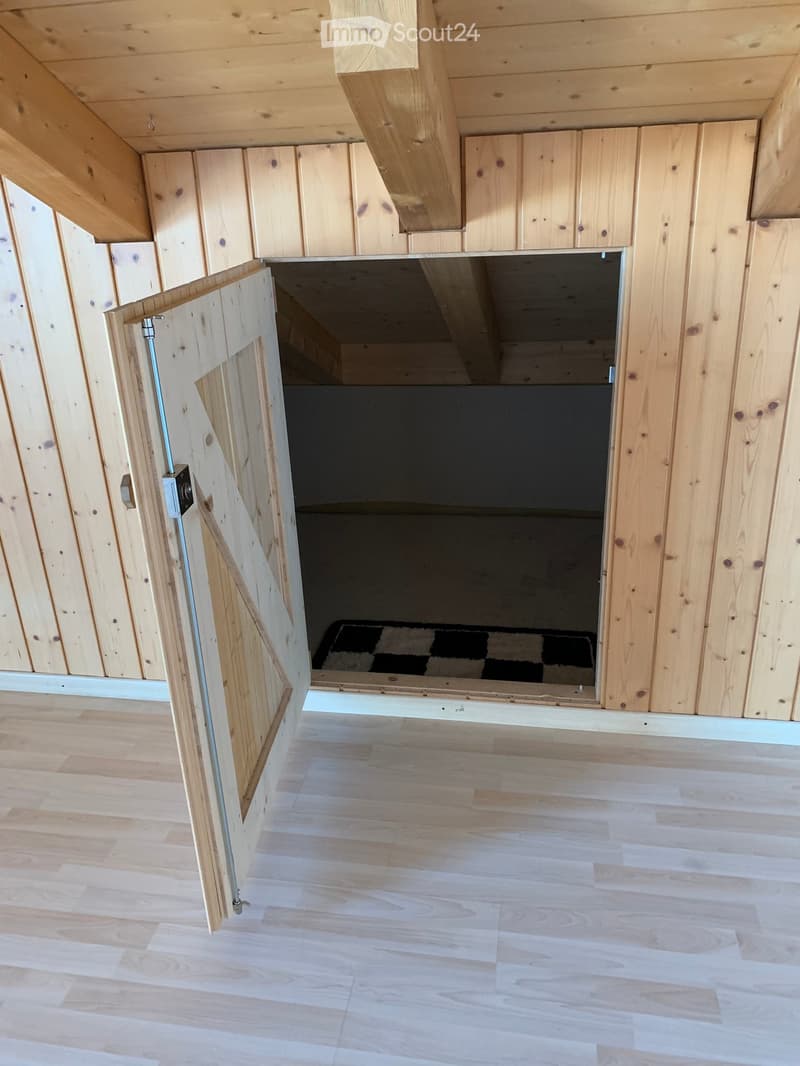 Empty room with light wooden walls and flooring, featuring an open doorway leading to another space with a dark floor and a small rug visible beyond.