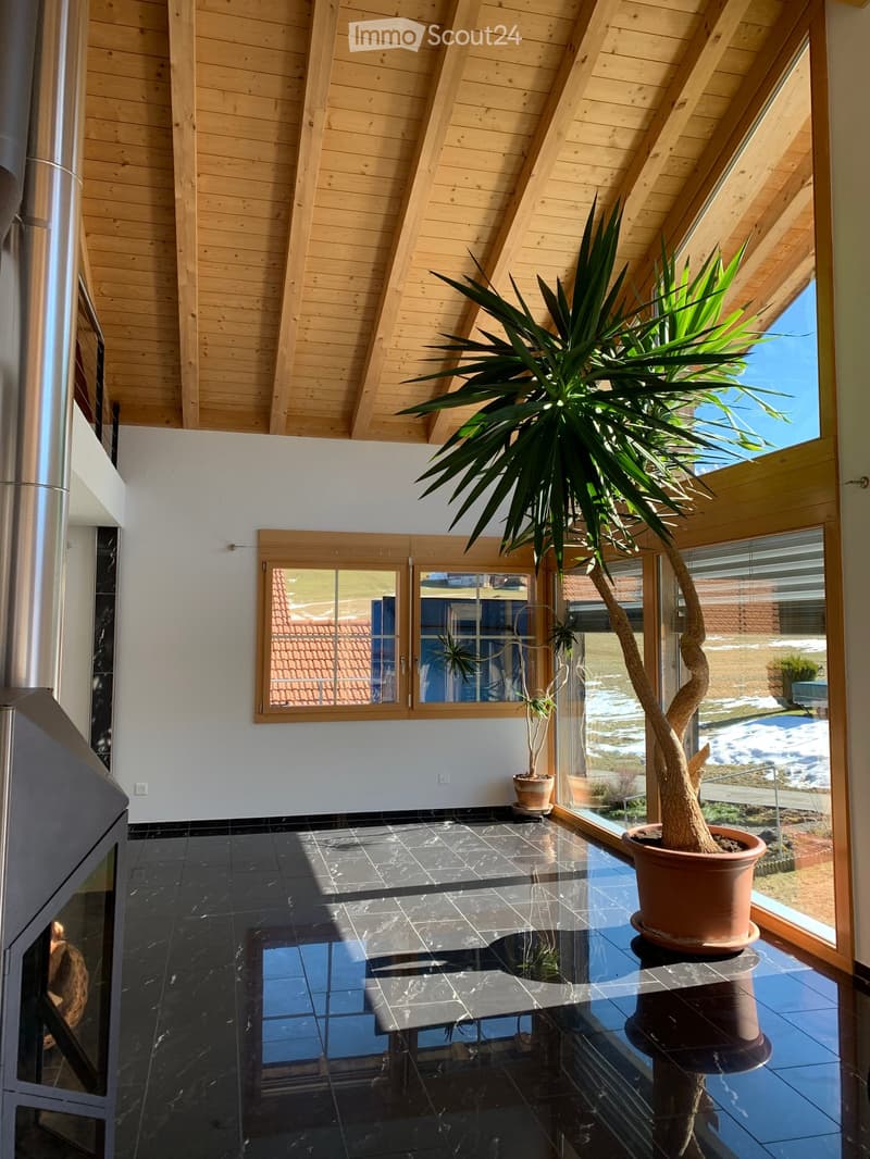 Bright, airy room with wooden ceiling beams, black tiled floor, large window with view, potted plant, and fireplace.