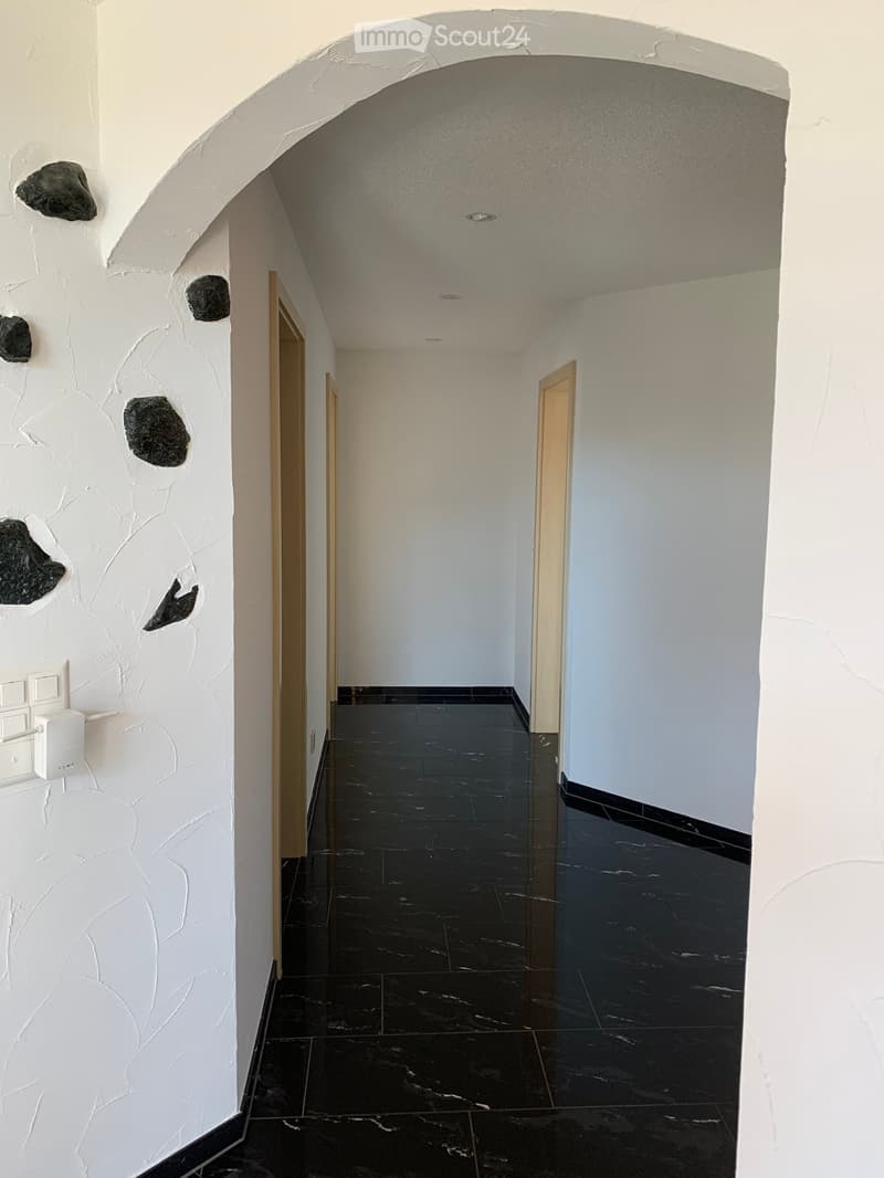 A modern hallway with black marble flooring, white textured walls, and recessed ceiling lighting. Features include a decorative stone accent on the left wall and wooden doors leading to other rooms.