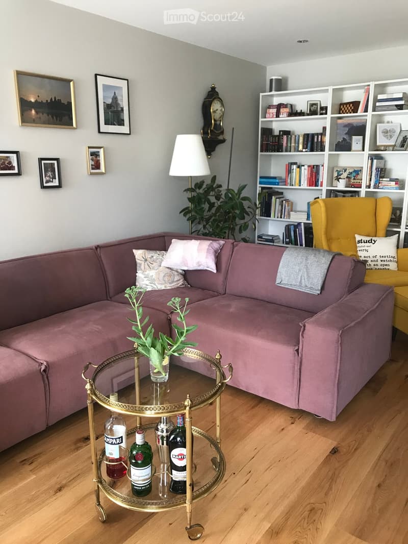 Living room with a pink sectional sofa, a gold-framed coffee table with bottles, a yellow armchair, a bookshelf, and a table lamp.