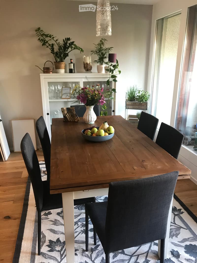 Dining room with a wooden dining table, six black chairs, a bowl of fruit, a vase with flowers, a bottle, potted plants, a cabinet, and a picture frame on the wall.