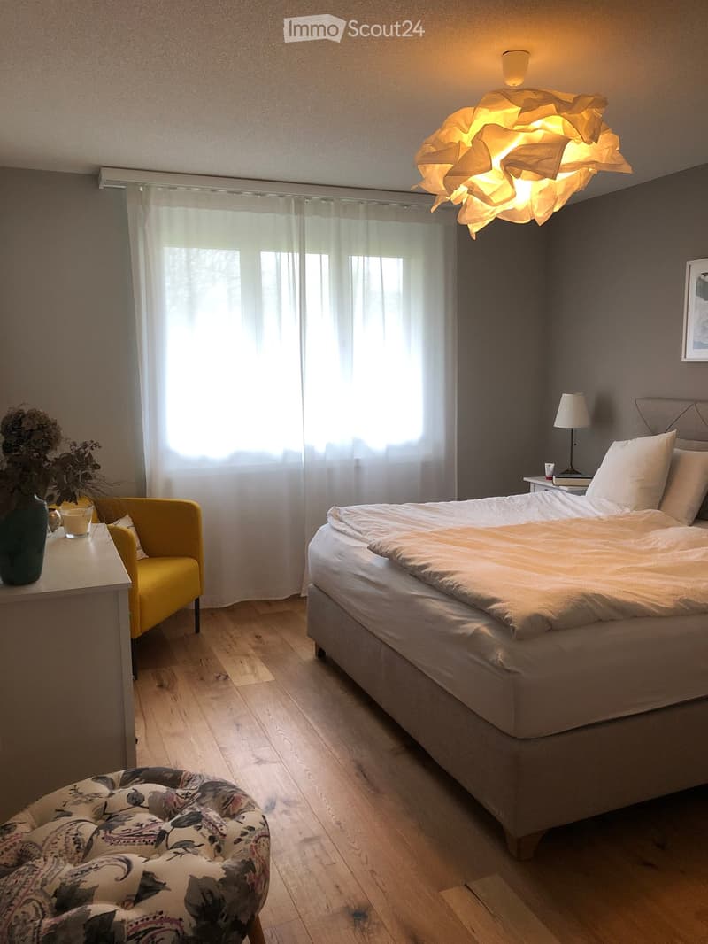 Modern bedroom with a large bed, wooden floor, large window with sheer curtains, yellow armchair, side table with lamp, decorative vase with flowers, and a patterned ottoman.