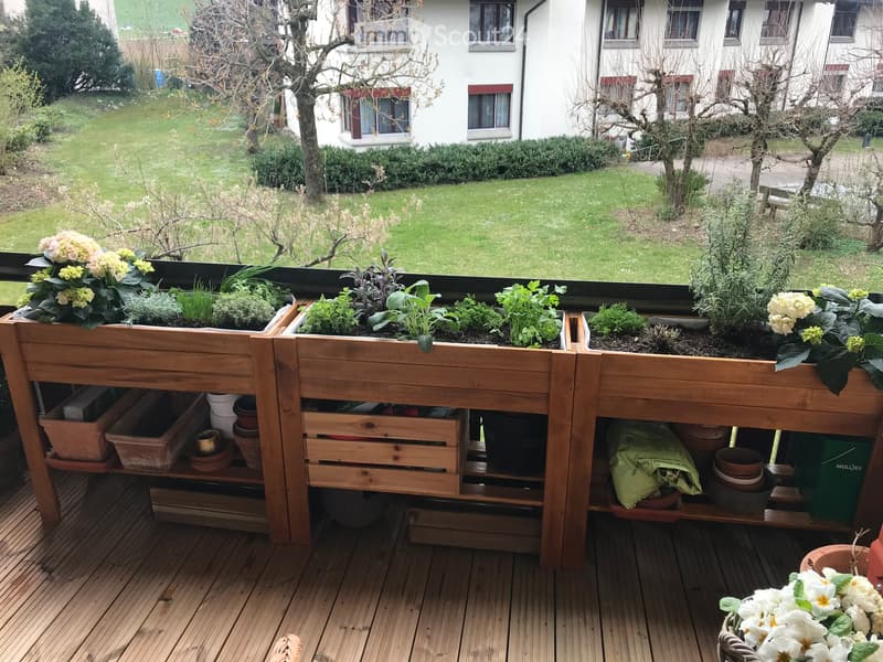 Balcony with wooden planters filled with herbs and flowers, wooden decking, and storage space underneath.