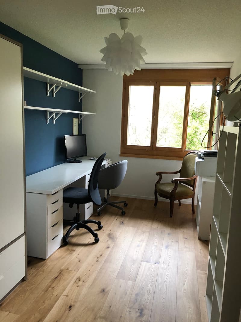 A modern, well-lit room with wooden flooring, a white desk, a black office chair, a computer screen, a wooden armchair, and built-in shelves.