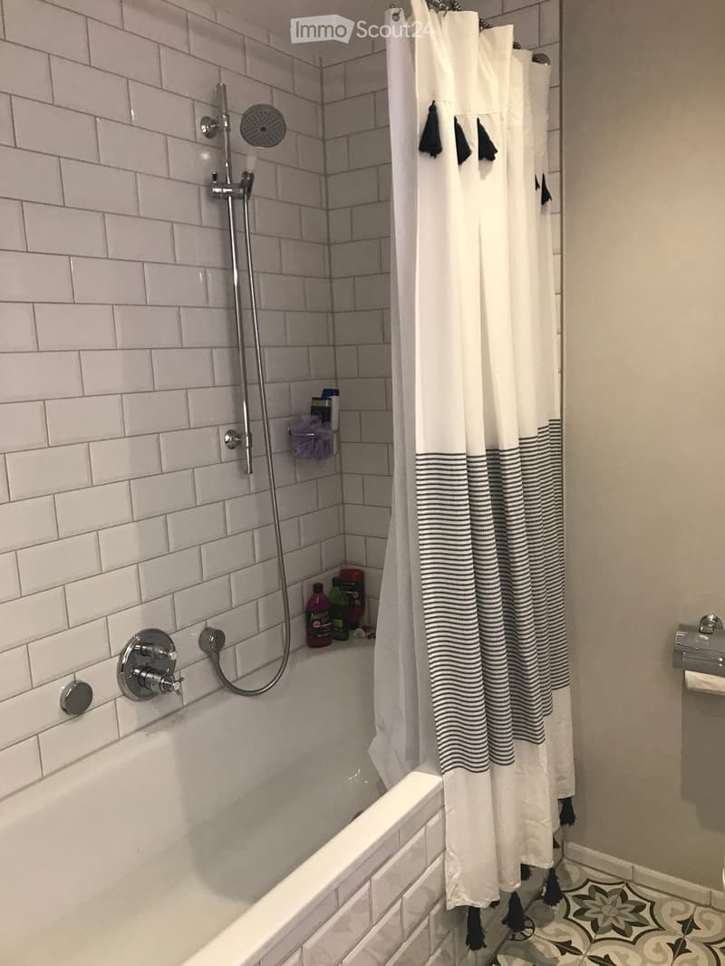 Modern bathroom with a white tiled shower area, bathtub, and decorative shower curtain. Includes a showerhead, faucet, and a few bottles on the edge of the tub.