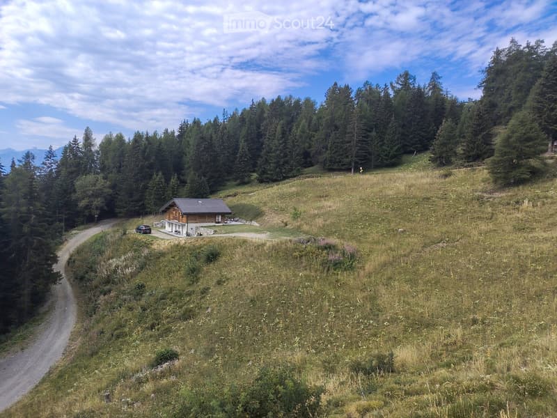 A small wooden house with a dark roof sits on a grassy hillside surrounded by dense forest and mountains under a partly cloudy sky.