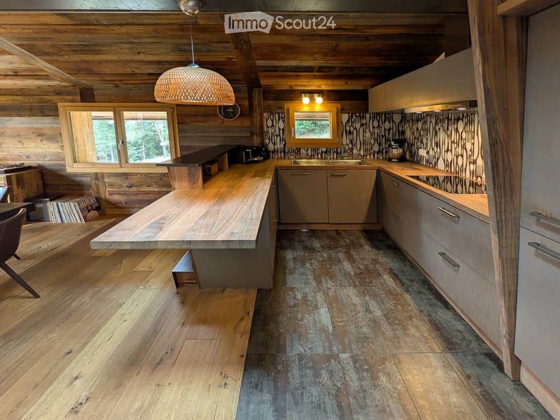 Modern kitchen with wooden beams, wooden countertops, and a mix of wooden and grey cabinets. Features a large island, sink, and a hanging pendant light.