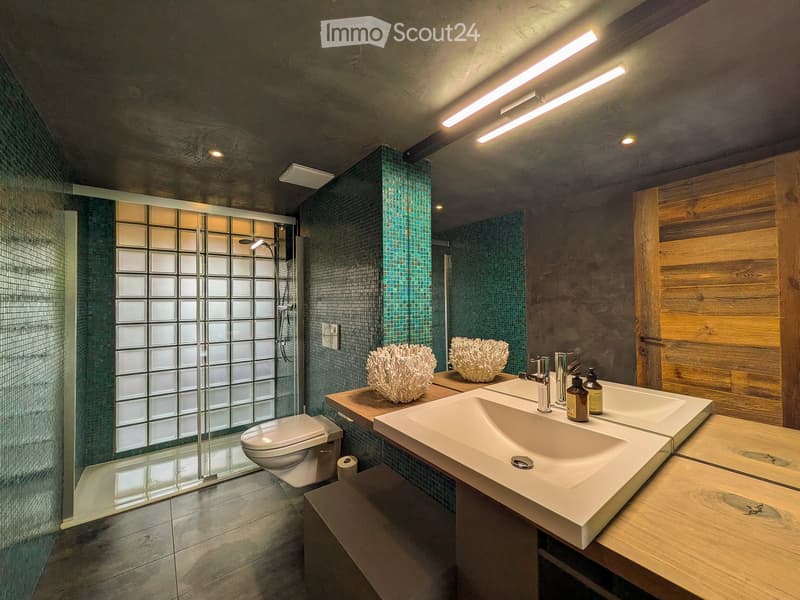 Modern bathroom with double sink, toilet, glass shower enclosure, decorative sculptures, and teal mosaic tiles.