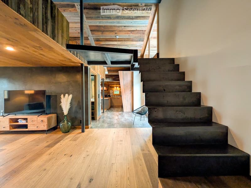Modern hallway with wooden floors, exposed wooden ceiling beams, and a staircase leading upwards. Features include a TV on a wooden stand, a vase with dried plants, and a view into a kitchen area with wooden cabinets and a dining table.