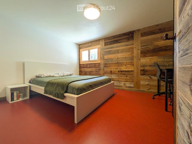 Modern bedroom with a white bed, green bedding, a nightstand with books, a wooden wall, and a small desk with a chair.