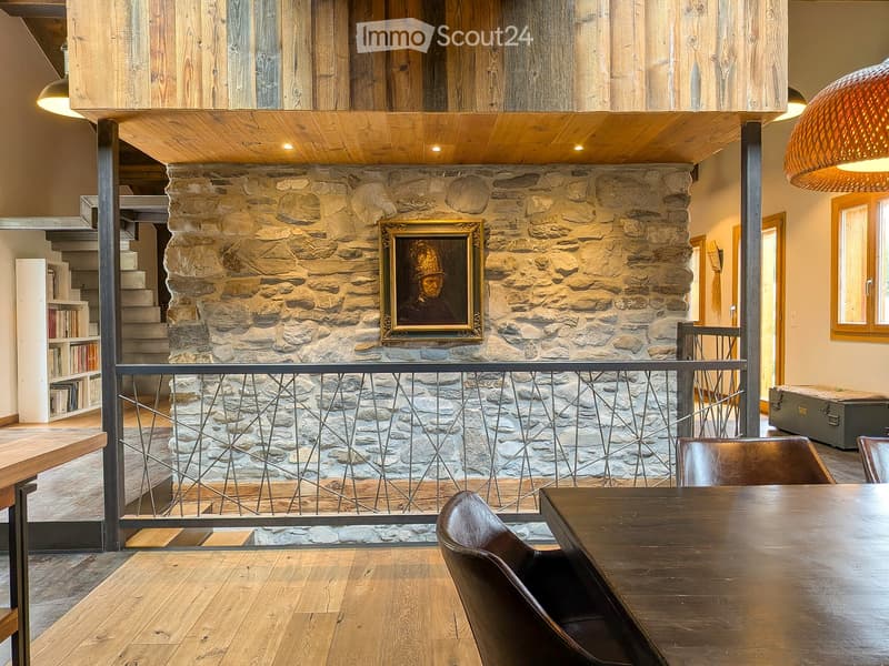 Spacious room with wooden floors, a stone wall, a large painting, a wooden table with leather chairs, a bookshelf, and a staircase leading to an upper level.