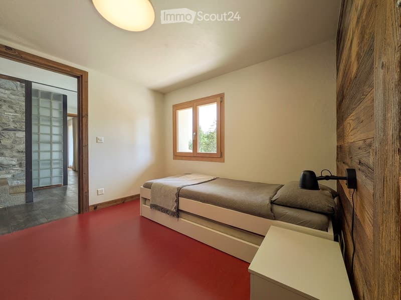 Bedroom with red floor, white walls, wooden window frame, bed with pillows and blanket, nightstand, and wall-mounted lamp.