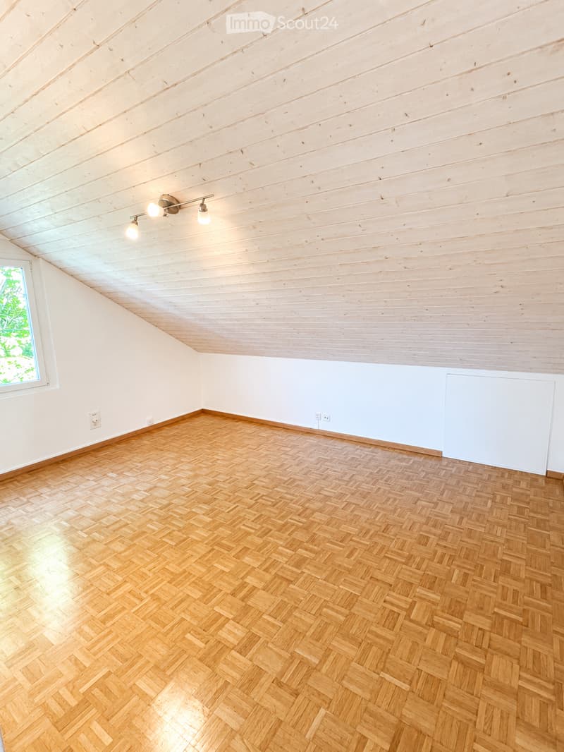 Empty room with wooden herringbone flooring, white walls, and exposed wooden ceiling beams. Features a single window and built-in lighting fixture.