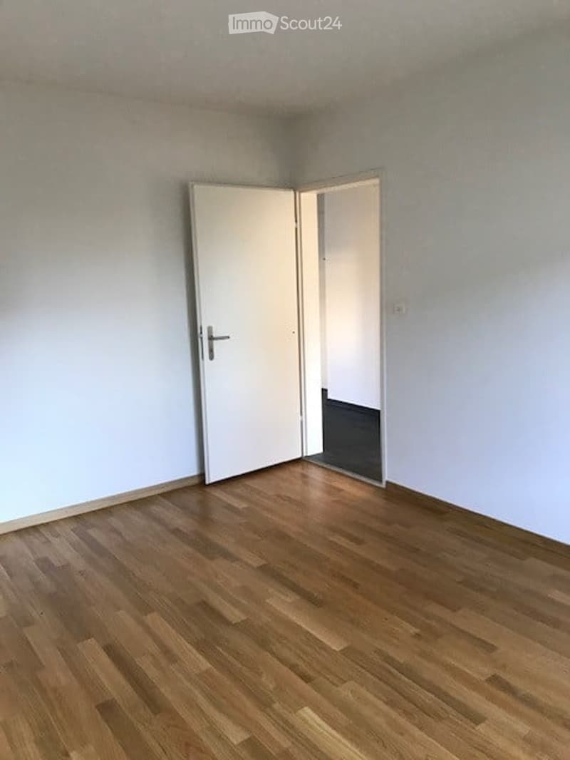 Empty room with light grey walls and wooden flooring, featuring an open white door leading to another room.