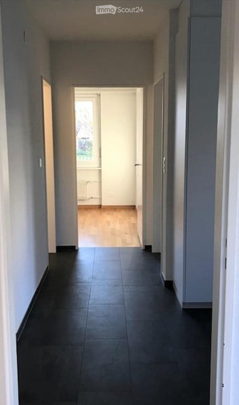 A long, narrow hallway with light grey walls, dark tiled flooring, and wooden flooring visible at the end of the corridor. The hallway leads to an open doorway with natural light coming through.