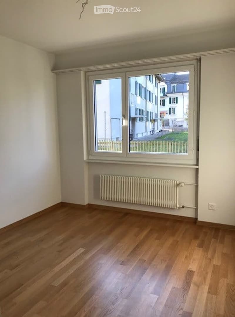 Empty room with wooden flooring, white walls, large window with white frame, radiator below the window, and visible outdoor area with a fence and buildings.