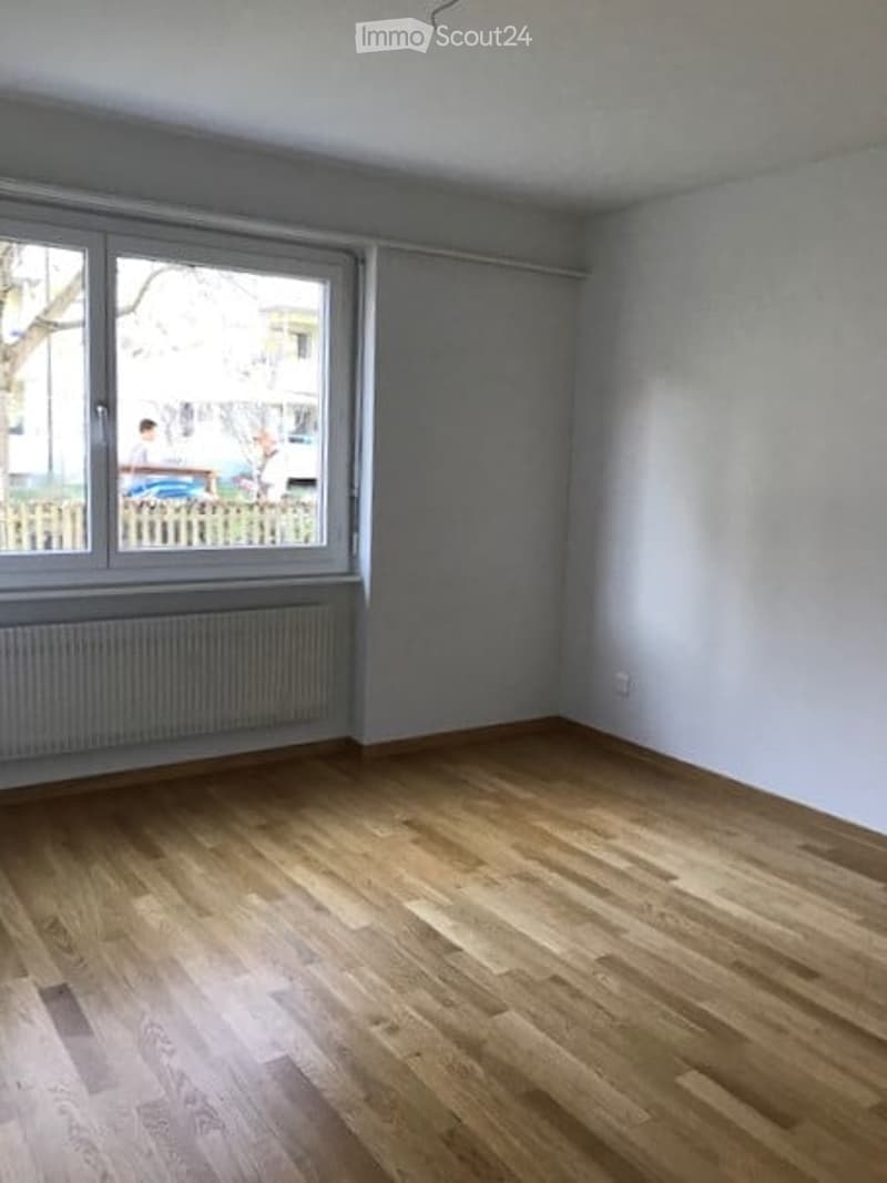 Empty room with wooden flooring, large window, radiator, and white walls.