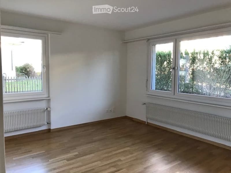 Empty room with wooden flooring, white walls, and large windows with white frames. Radiators are present under the windows on both sides.
