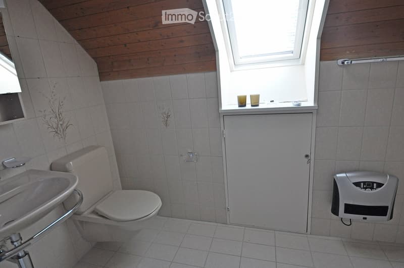 Bathroom with a toilet, sink, hand dryer, and a small window with two cups on the sill. The walls are tiled, and the ceiling is wooden.