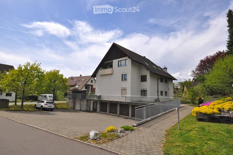 Two-story house with white exterior, dark roof, and balcony on the upper level. The house is surrounded by a well-maintained garden with colorful flowers and trees. There is a paved driveway leading to a garage area with multiple parking spaces.