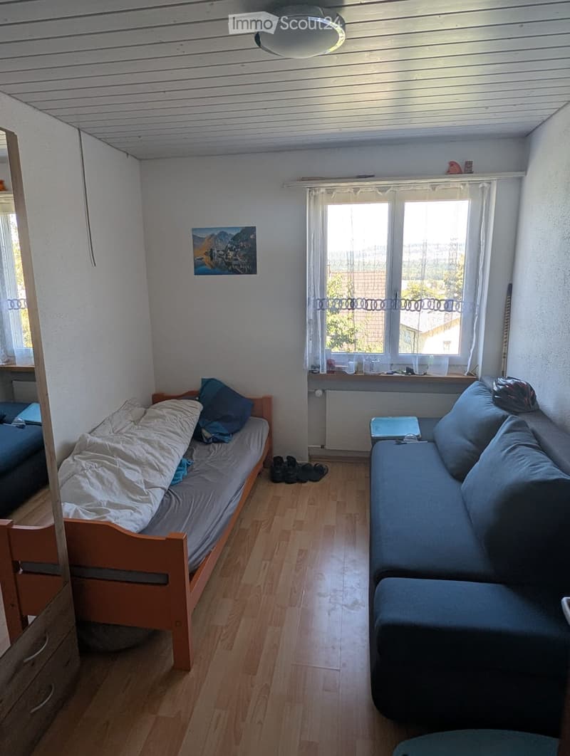 A small, cozy room with a single bed, a sofa, a small table, and a picture frame on the wall. The room has wooden flooring and large windows with white curtains.