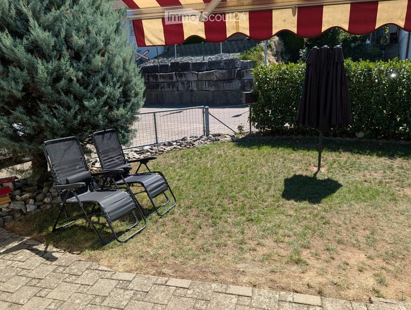 Garden area with two black folding chairs, a closed umbrella, and a striped awning in the background.