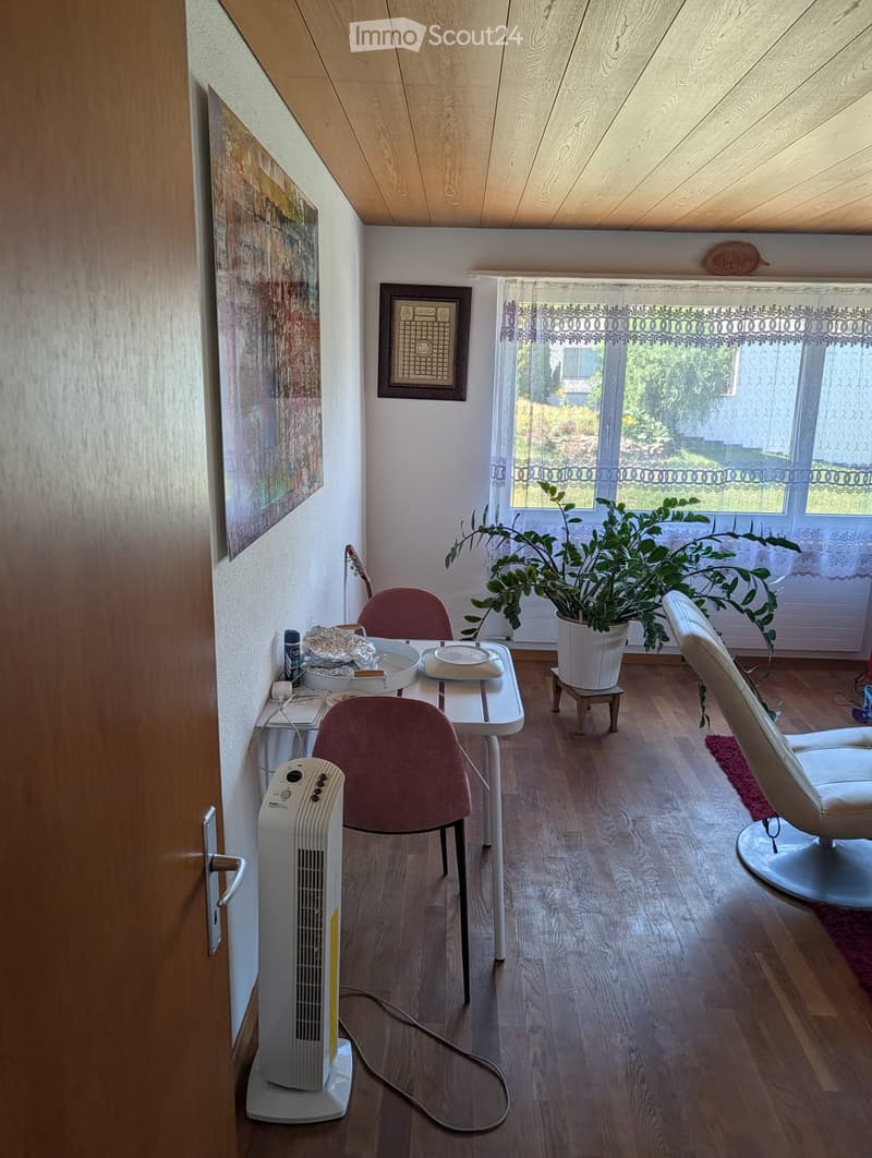 A cozy room with wooden flooring, a white table with chairs, a fan, a large potted plant, and a white leather chair near the window.