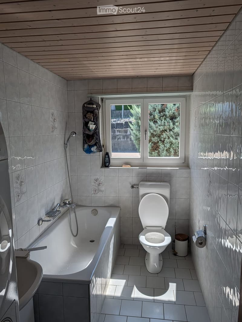 Modern bathroom with a bathtub, toilet, and shower hose. Features tiled walls and floor, a window with a view of greenery, and a small shelf holding toiletries.
