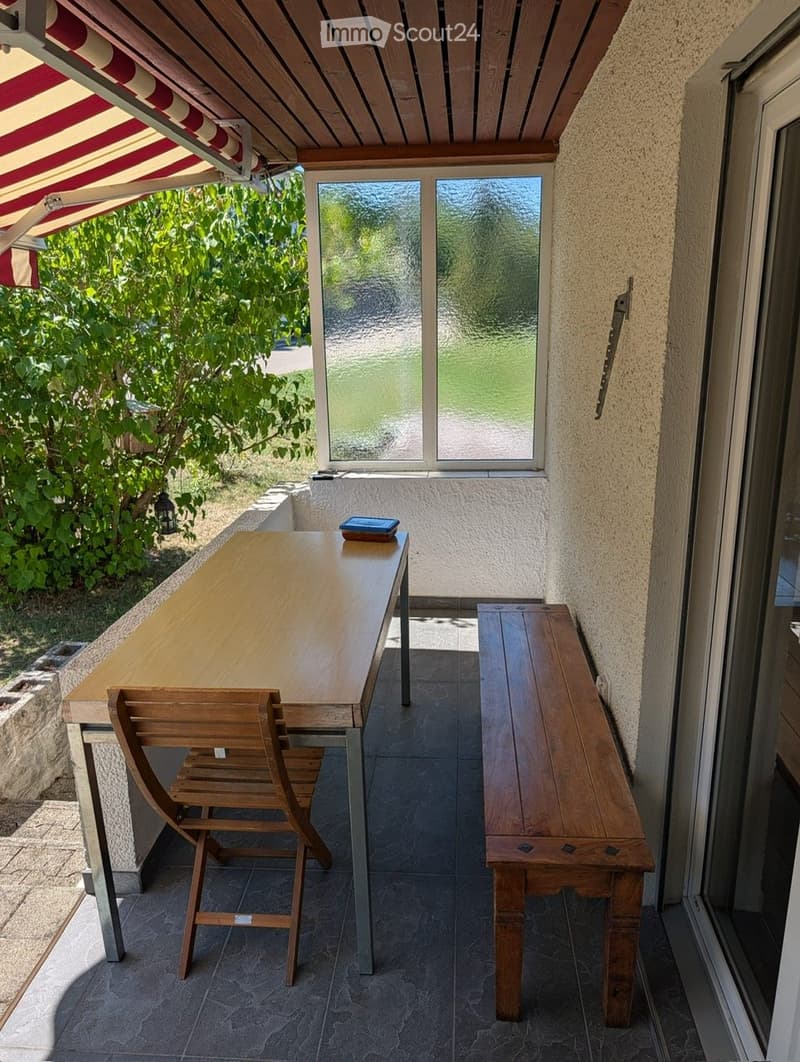 Balcony with a wooden table, a wooden chair, and a wooden bench, featuring a striped awning and a view of greenery.