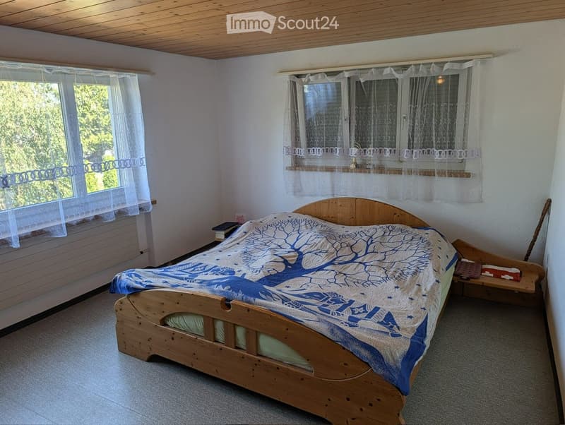 Cozy bedroom with a wooden bed, blue and white patterned bedspread, nightstand with books, and large windows with sheer curtains.