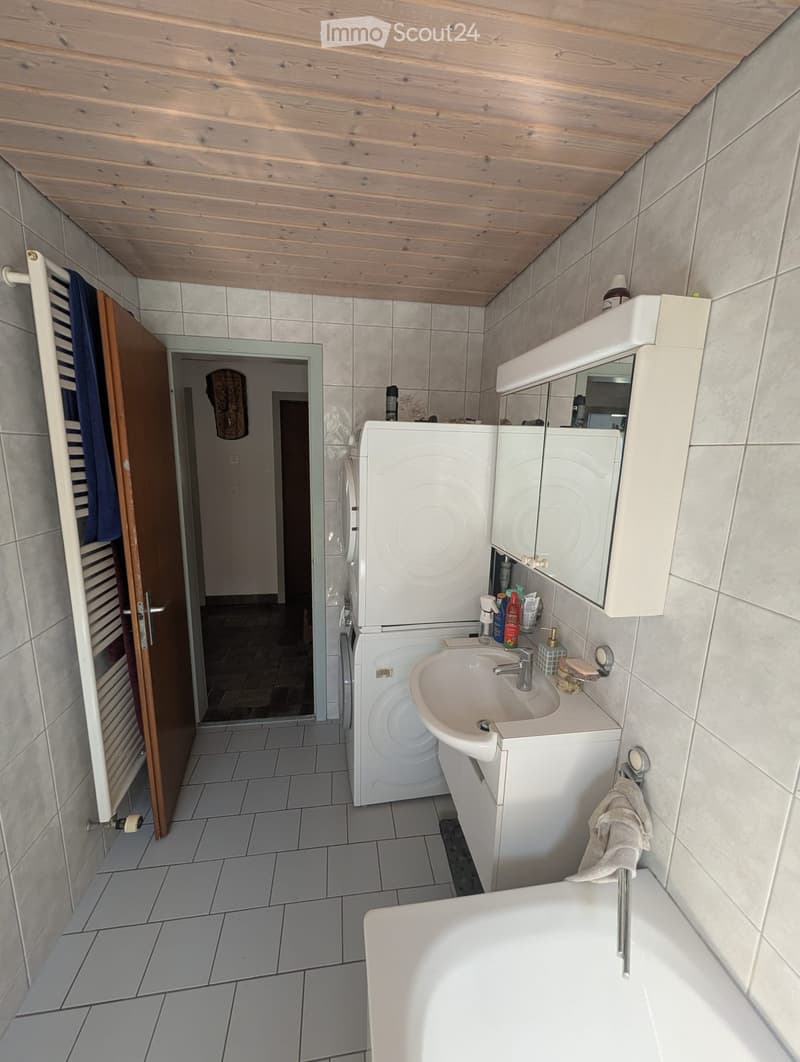 Bathroom with a sink, bathtub, mirror, cabinet, and washing machine. The room has tiled walls and floor, a wooden door, and a towel hanging on a rack.