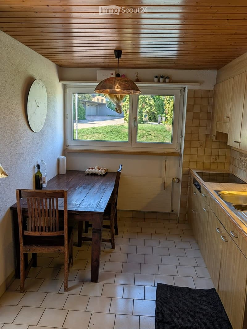 Bright kitchen with wooden ceiling, tiled floor, and large window overlooking a green lawn. Includes a wooden dining table with chairs, a bottle on the table, a clock on the wall, and modern cabinets with a sink.