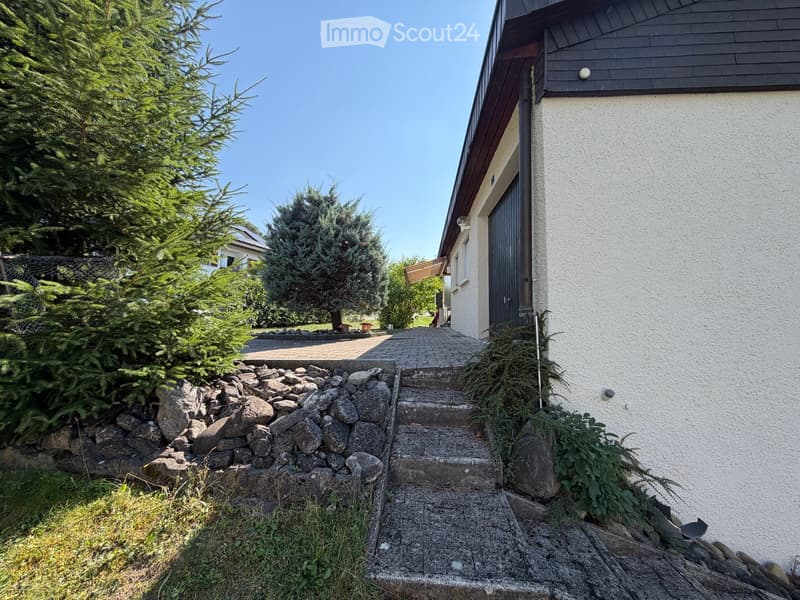 Garden area with stone steps leading to a house entrance, surrounded by greenery and rocks.