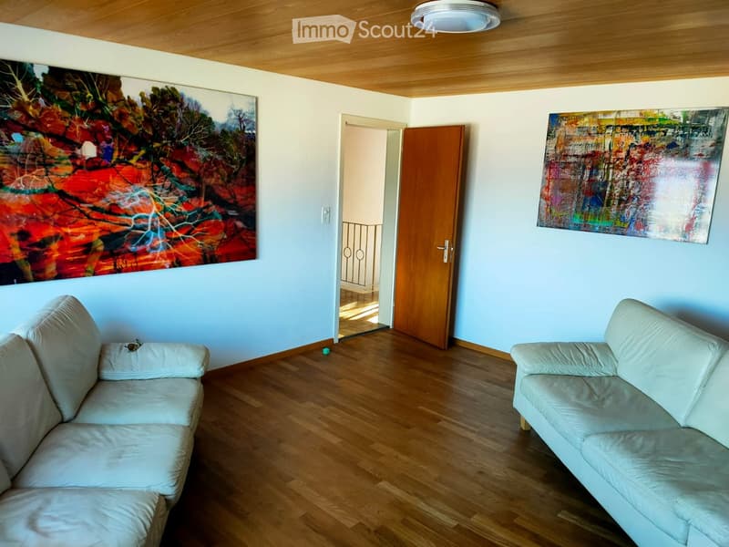 Living room with light-colored sofas, wooden flooring, and two large abstract paintings on the walls. A wooden door leads to a balcony area.