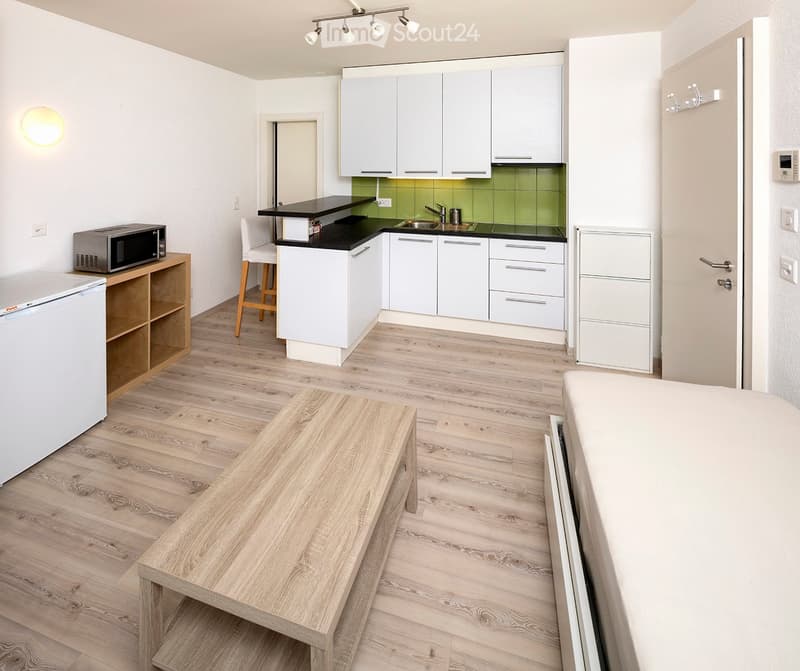 Modern, open-plan living and kitchen area with light wooden flooring, white kitchen cabinets, a small dining table, a microwave, and a refrigerator.