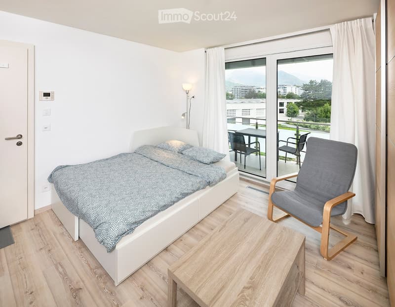 Cozy bedroom with a single bed, a wooden chair, a small table, and a view of the mountains through large windows and a sliding glass door leading to a balcony.