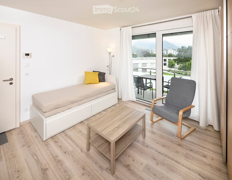 Modern furnished room with a sofa, small table, chair, and lamp, featuring large windows with a scenic mountain view.